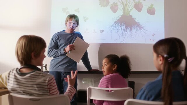 Rear view of three intercultural youthful learners sitting on chairs in front of teacher making report or presentation on interactive screen while clever schoolboy explaining one of points