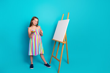 Smiling young girl in a colorful striped dress with painting tools standing next to a blank canvas...