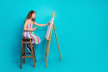 Young girl in colorful striped dress painting on canvas while sitting on stool with a joyful summer...