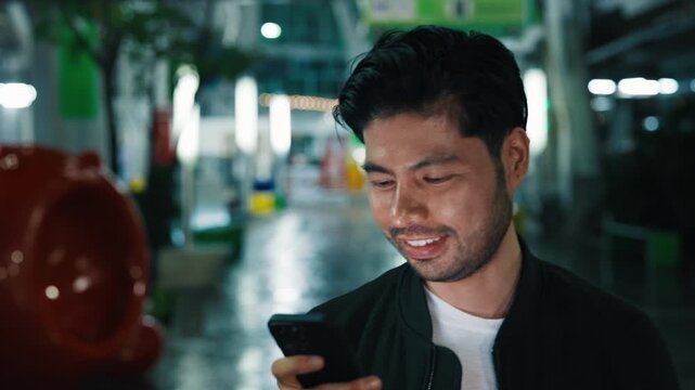 Portrait of cheerful male with fancy hairdo walking by downtown district while looking at screen of cellular phone. Brunette man scrolling social media and observing new trends in comedy videos.
