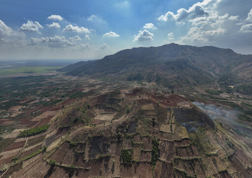 Aerial view of terraced fields cascade down the rugged slopes of a mountain, a patchwork of earthy browns and greens against a backdrop of distant plains, Chu Pah, Gia Lai, Vietnam.