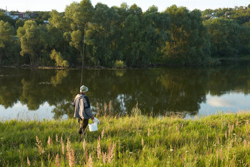 A lone fisherman with a fishing rod and a bucket in his hands goes down to a picturesque pond...