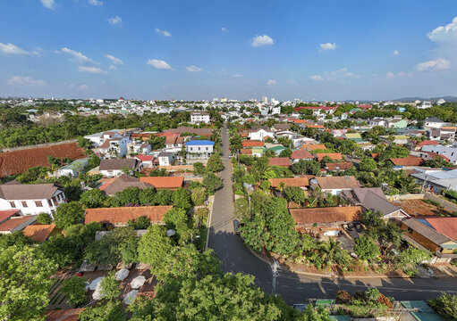 Aerial view of terracotta roofs nestled among verdant trees, bisected by a smooth grey road under a bright sky, Buon Ma Thuot, Dak Lak, Vietnam.