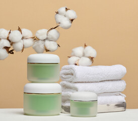Green cosmetic jars, stack of white towels and branches of natural cotton are arranged on a beige...