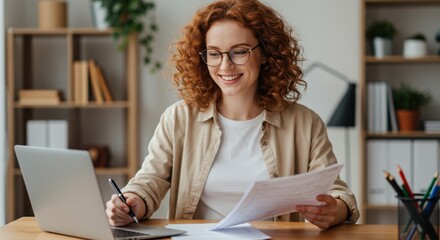 Smiling red haired woman wearing glasses works on laptop and reviews documents at her desk