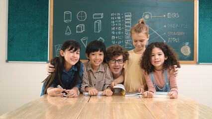 Caucasian professional teacher sitting while hugging diverse cute student at class and looking at camera. Group of smart multicultural children smiling to camera while standing at classroom. Pedagogy.