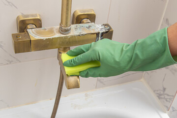Man or woman in green gloves cleans a shower faucet from plaque with a sponge. Cleaning the bathroom