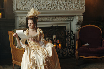 Caucasian young adult woman wearing elaborate Baroque style dress sitting in ornate chair reading...