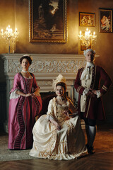 Three Caucasian adults in Baroque period costumes posing in ornate Renaissance style interior, two...