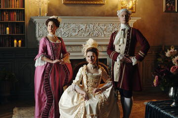 Three Caucasian adults in Baroque period costumes posing in ornate Renaissance style interior, two...