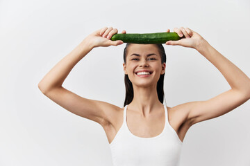 Smiling young woman holding cucumber over her head, bright and cheerful expression in simple, clean...