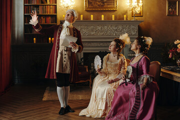 Caucasian middle aged man in Baroque costume gesturing dramatically while holding paper, two...