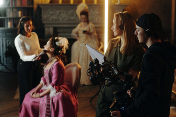 Caucasian young adult woman sitting in chair receiving makeup from another woman while film crew...