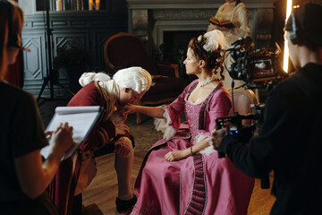 Caucasian middle aged man in Baroque costume kneeling and kissing hand of young woman in Renaissance dress while film crew recording scene