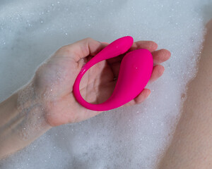 A woman lies in a bubble bath and holds a curved pink vibrator.