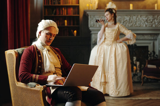 Caucasian middle aged man in Baroque period costume sitting on chair using laptop against woman in Renaissance style dress