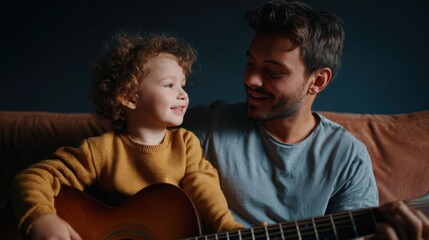 Father and his young son sitting on a brown couch. the father is holding an acoustic guitar and the son is looking up at him with a smile on his face.