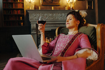 Middle aged Caucasian woman reclining in ornate chair holding wine glass and using laptop, wearing...