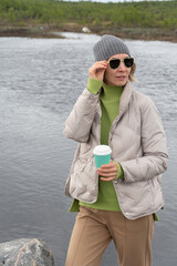 Woman in a beige puffer jacket and gray beanie stands by an Arctic lake holding a turquoise...