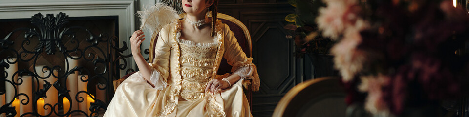 Caucasian young adult woman sitting in ornate chair holding feather fan wearing elaborate Baroque...
