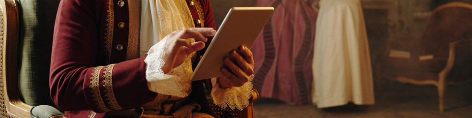 Caucasian middle aged man wearing Baroque style costume using digital tablet while sitting indoors,...