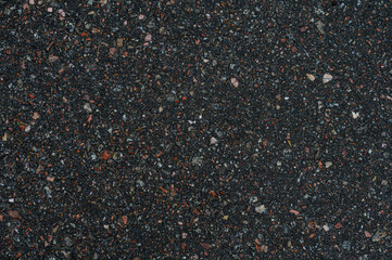 Close up view of dark colored gravel with small, multicolored stones. The texture is coarse and granular, showing a mix of dark gray, black, and small specks of red and other colors.