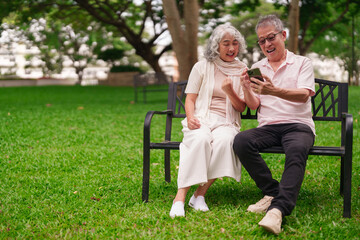 Asian elderly couple happy using mobile phone app park bench leisure outdoor activity after retirement fun senior life digital technology