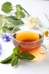 Aromatic herbal tea in glass cup, different flowers and mint on white background, closeup
