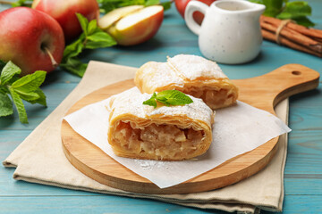 Pieces of tasty apple strudel with powdered sugar, mint, fruits and cinnamon on light blue wooden...