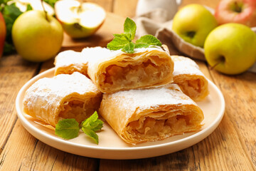 Pieces of tasty apple strudel with powdered sugar, mint and fruits on wooden table, closeup