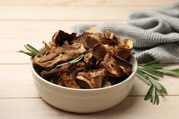 Pieces of dried mushrooms and rosemary in bowl on light wooden table, closeup