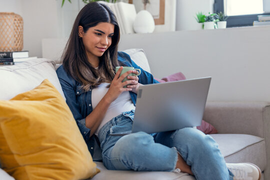 Young woman working with her laptop while drinking a cup of coffee sitting on a couch at home.