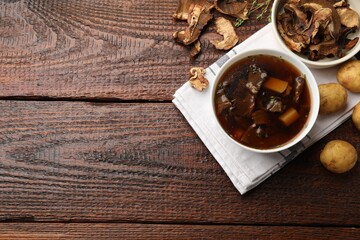 Tasty soup in bowl, raw potato and dry chanterelle mushrooms on wooden table, flat lay. Space for text