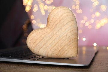 Online dating site. Wooden heart and laptop on table against blurred lights, closeup. Bokeh effect