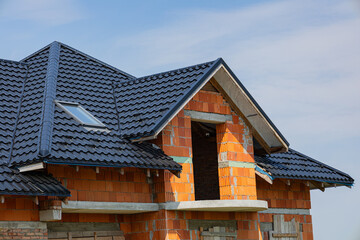 Metal tile roofing is being installed on a partially built house under a clear blue sky, showcasing modern construction techniques