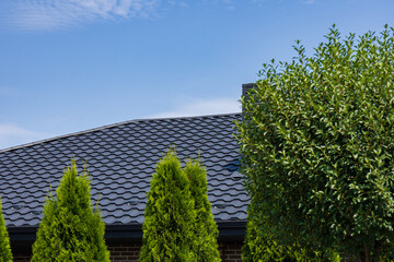 A sleek metal tile roof showcases its design atop a brick residence, surrounded by lush green trees under bright blue skies