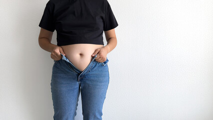 Closeup of woman struggling to zip up tight jeans against white wall, visible belly, concept of...
