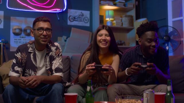 Front view shot of multiethnic group of friends playing videogames sitting on sofa and holding controllers having fun together