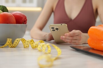 Woman using smartphone at white marble table with vegetables, kitchen scale and measuring tape...