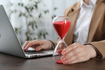 Deadline. Woman with hourglass and laptop working at wooden table in office, closeup