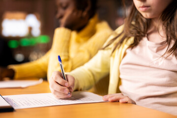 Two diverse schoolgirls focused on writing an essay in a school library, completing their school...