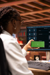 Woman using mockup smartphone and AI LLM chatbot on computer screen in home office. African...