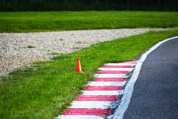 Close-up view of a racing track corner with red and white curb markings and an orange traffic cone on green grass. Ideal for illustrating concepts of motorsport safety, outdoor racing