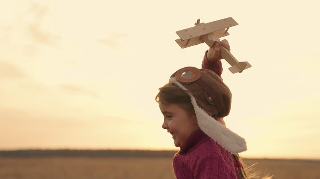 little child runs with toy airplane in his hand at sunset, childhood dream of flying, kid plays in pilot in helmet, little baby outdoor activities, wooden toy cheerful girl traveling rays of sun light