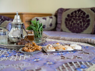Traditional Moroccan tea service with mint leaves dates and assorted sweets on ornate glass tray with decorative cushions