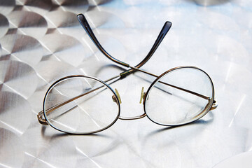 A pair of round metal eyeglasses with thin frame rests on a patterned metallic cafe table. the...