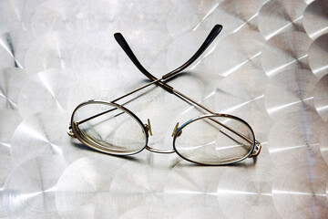 A pair of round metal eyeglasses with thin frame rests on a patterned metallic cafe table. the vintage style spectacles lie in natural light, creating a soft reflective effect. 
