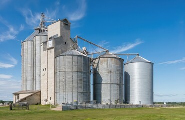 Industrial landscape of concrete grain silos under clear blue sky. Gray silos with metal roofs in two rows of three. Vast grassy field surrounds silos, with fence on left side. Perspective gives