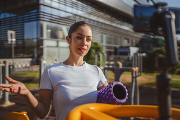 sportswoman at outdoor gym filming herself with foam roller on camera