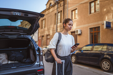 woman with suitcase use cellphone in front car trunk on the street